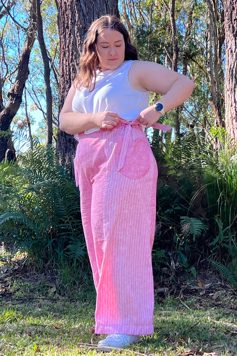 A woman standing outdoors in a wooded area wearing a white sleeveless top and pink vertically striped wide-leg pants, adjusting the waist tie of the pants.