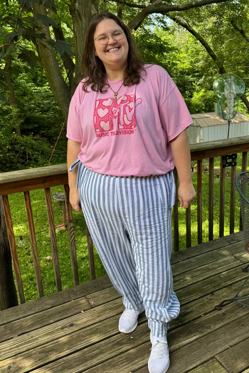 A woman stands on a wooden deck outdoors wearing a pink graphic t-shirt, blue and white striped pants, and white sneakers.