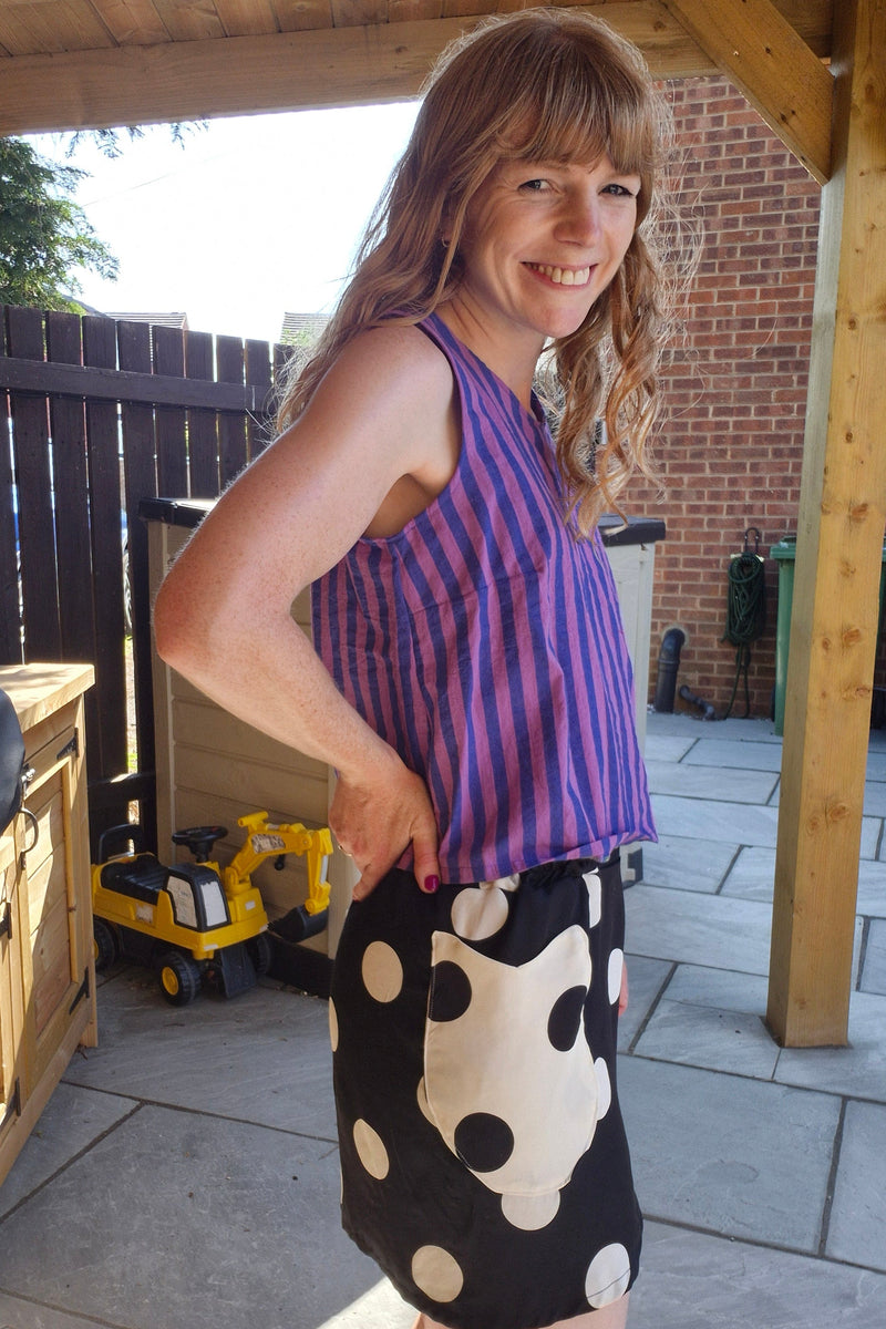 A woman wearing a purple and blue striped sleeveless top and a black skirt with large white polka dots, standing outdoors on a paved patio under a wooden structure.