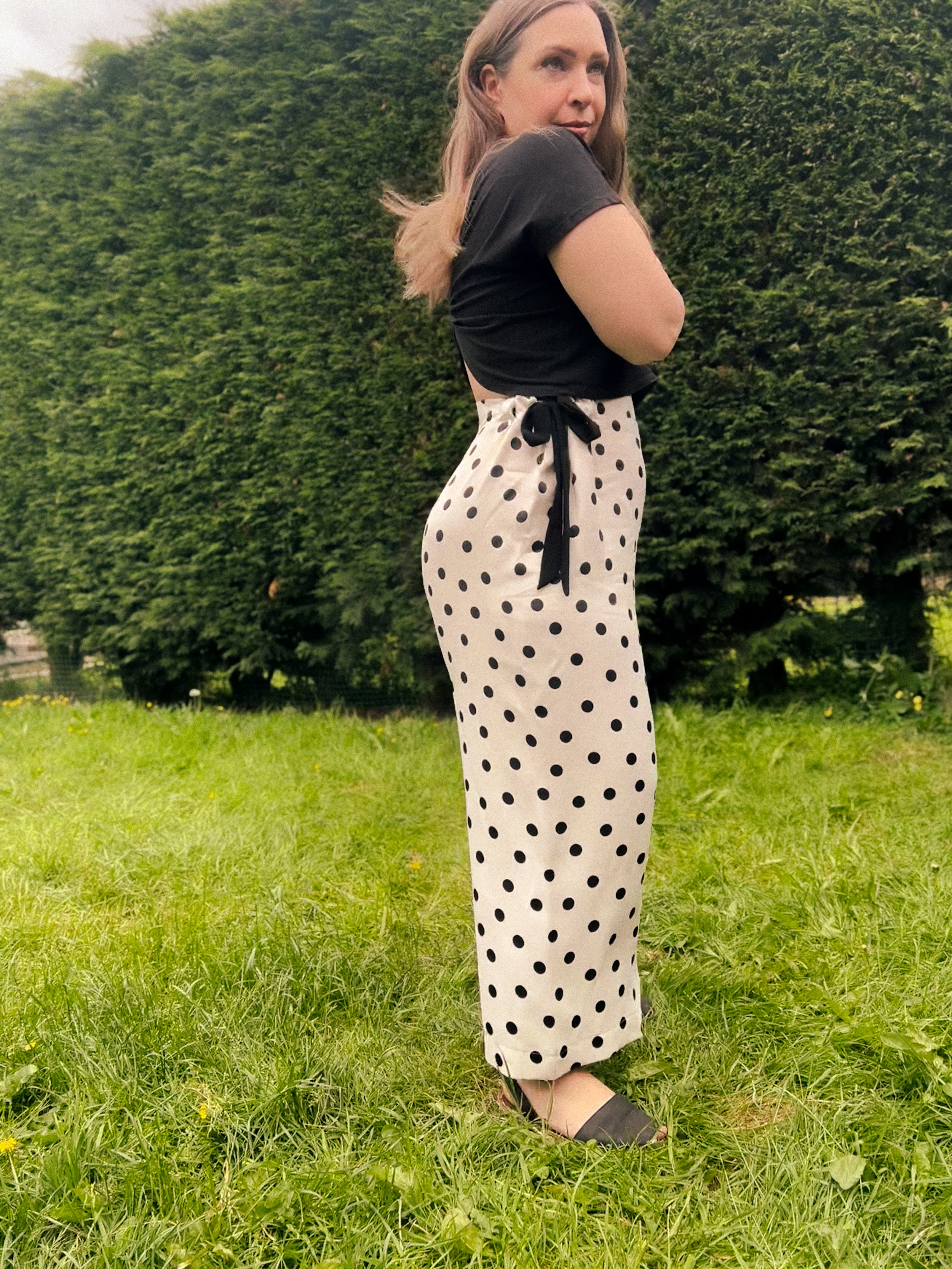 A woman standing on a grassy lawn in front of a green hedge, wearing a black short-sleeve crop top and cream-colored wide-leg trousers with black polka dots and a black waist tie.