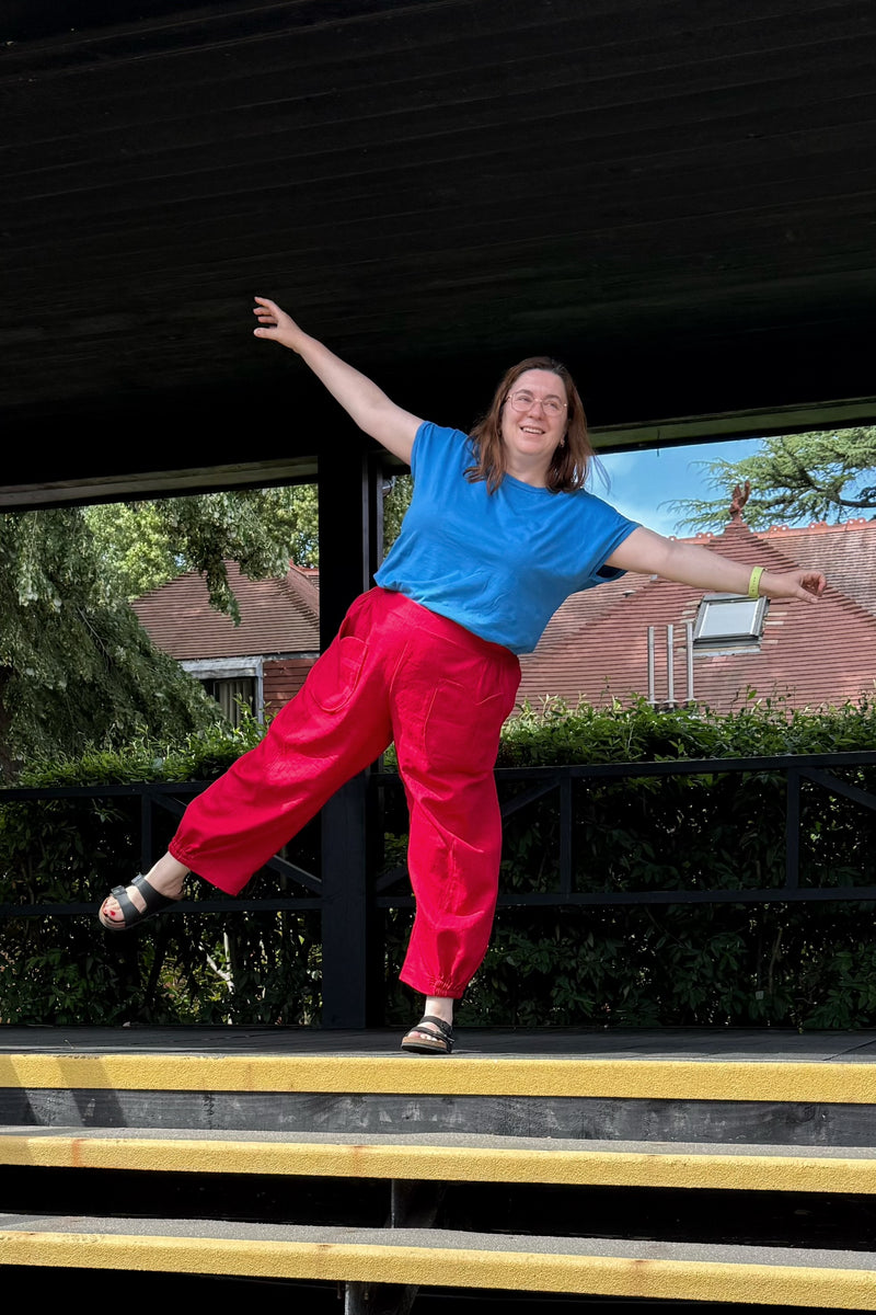 A woman posing on outdoor wooden steps under a dark shelter, wearing a bright blue t-shirt and vibrant red wide-leg pants.
