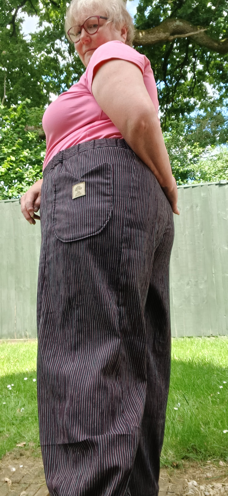A person outdoors wearing dark charcoal pinstriped overalls with a pink shirt, photographed from a low angle with trees and grass in the background.