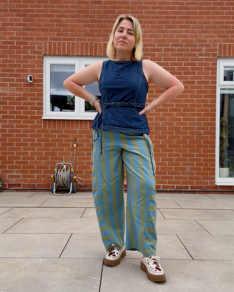 A woman stands outdoors in front of a red brick wall, wearing a sleeveless dark blue denim top with a braided waist detail and wide-leg striped pants in blue and olive green, paired with brown platform sneakers.