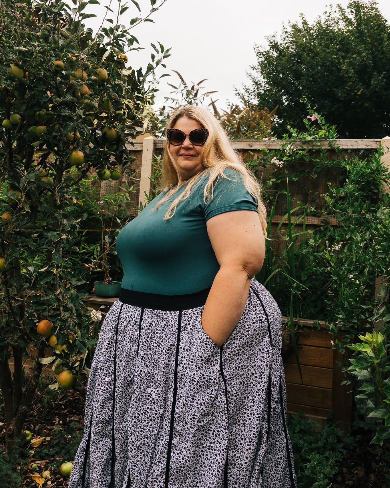 Woman in a green top and patterned skirt standing in an outdoor setting with trees and plants.