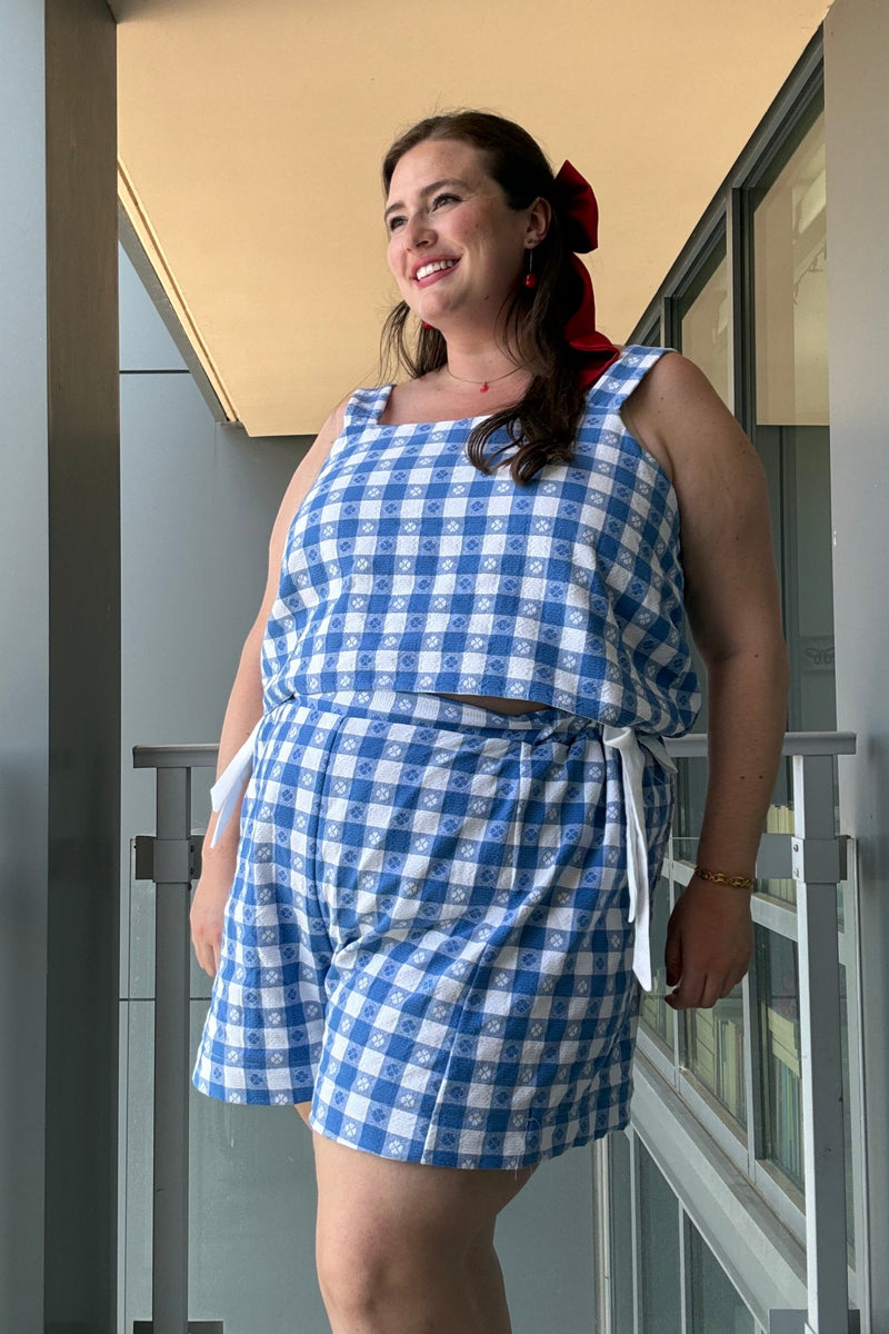 A woman wearing a blue and white gingham check romper stands on a modern balcony with glass railings and a beige ceiling.