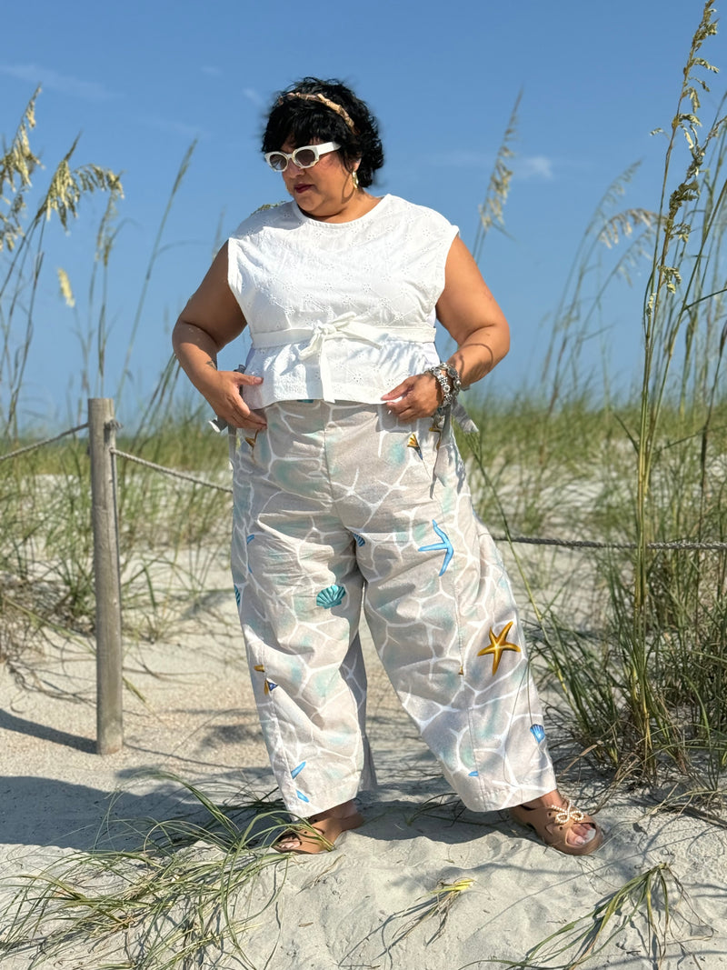 A woman stands on a sandy beach wearing a white sleeveless top and white wide-leg pants with blue and yellow starfish and shell prints, set against beach grass and blue sky.