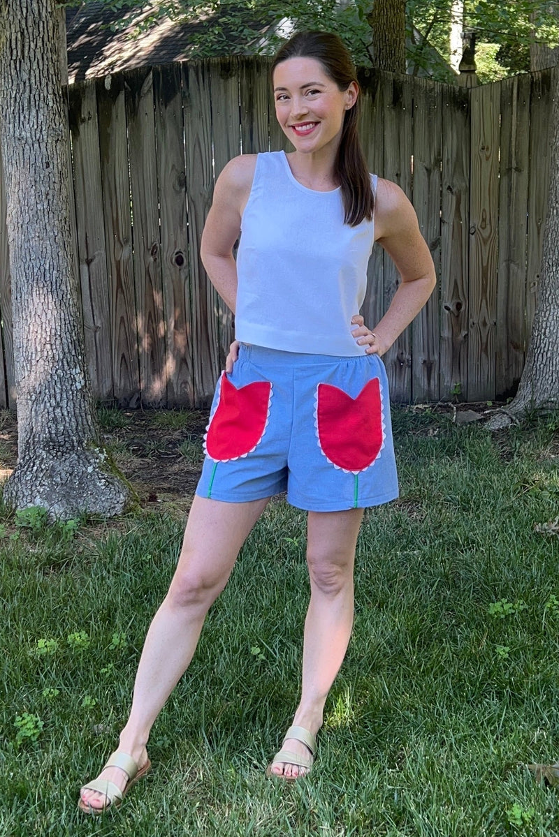 A woman standing outdoors on grass in front of a wooden fence, wearing light blue shorts with large red patch pockets and a white sleeveless top.
