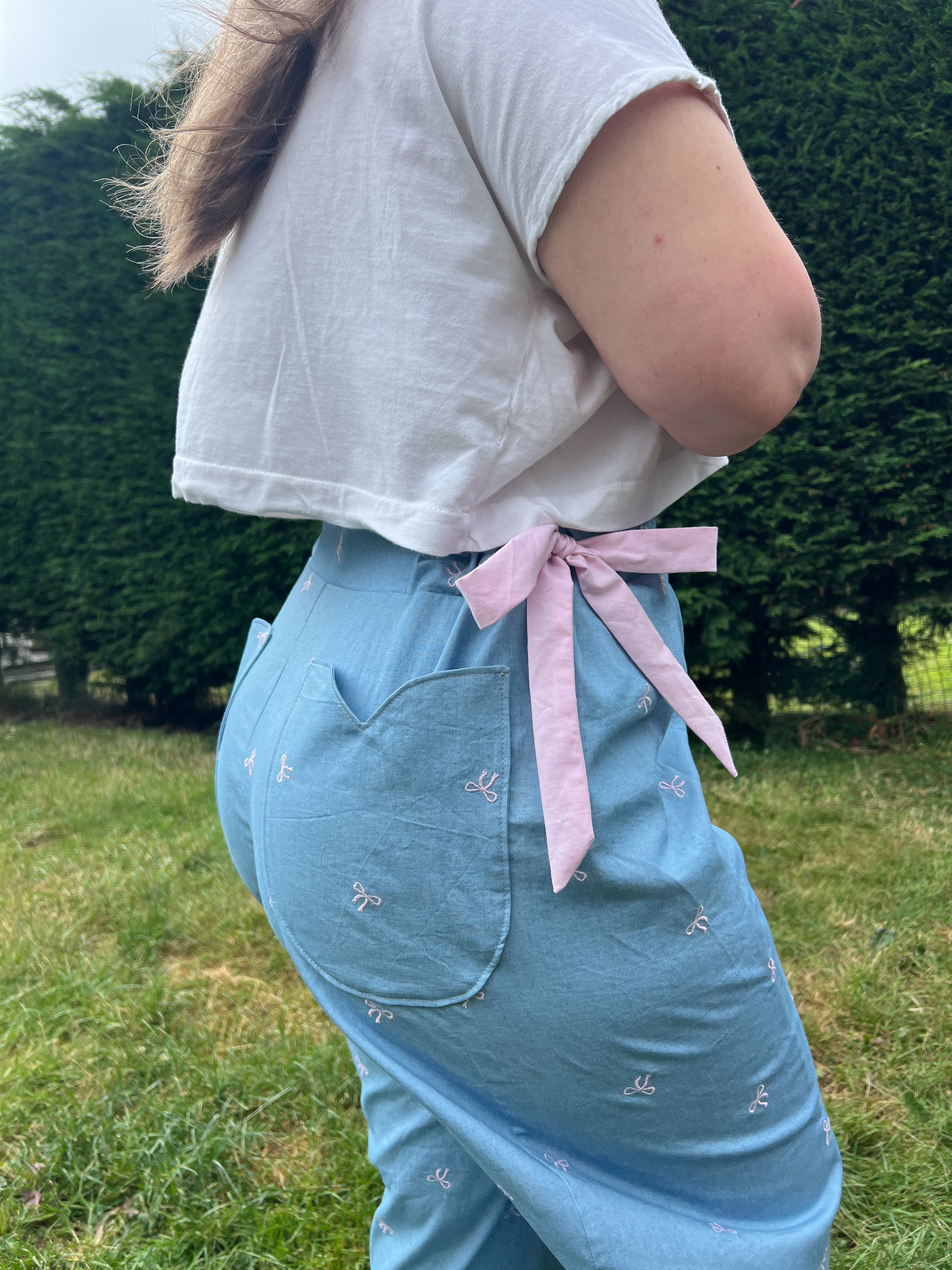 A person outdoors wearing light blue embroidered pants with a pink ribbon tied at the back waist, paired with a white cropped T-shirt.