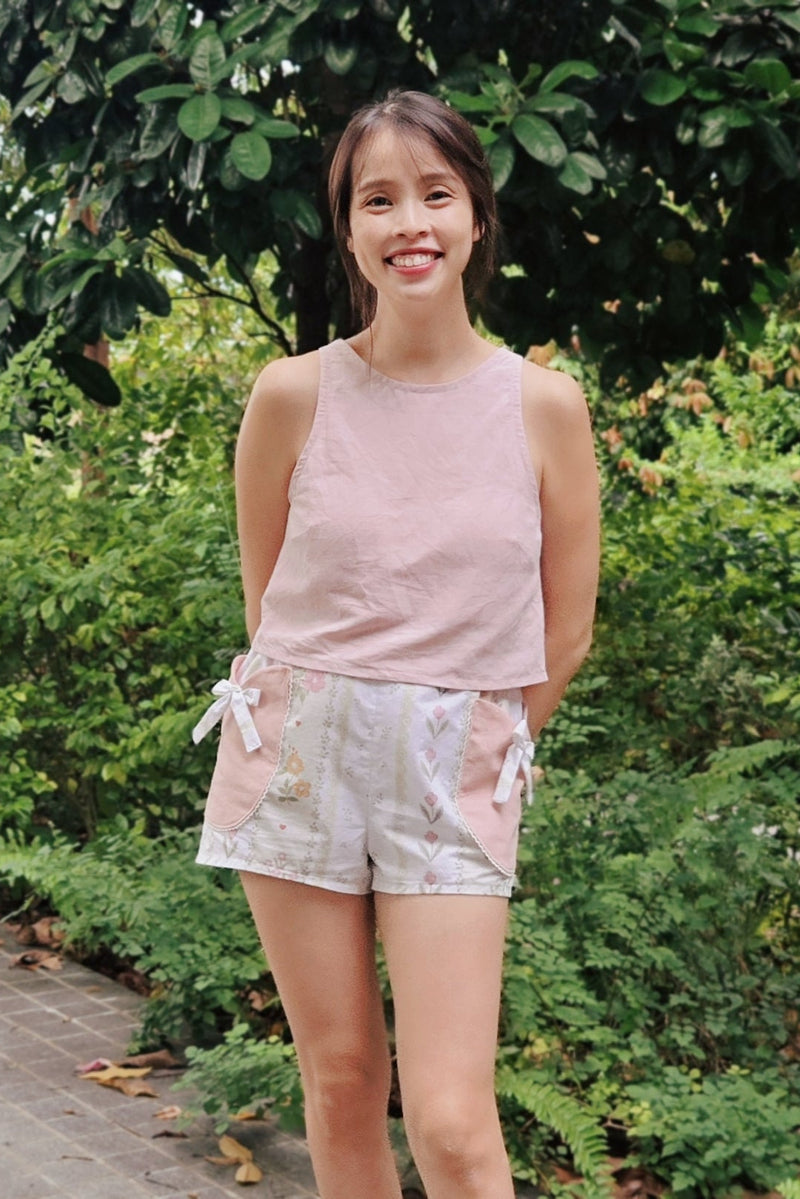 A woman standing outdoors in front of green foliage, wearing a light pink sleeveless top and light patterned shorts with white tie details.