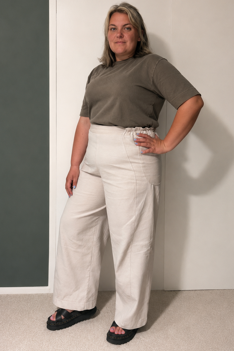 A woman modeling a short-sleeve olive green t-shirt and high-waisted, wide-leg off-white trousers, standing against a split white and dark gray wall.