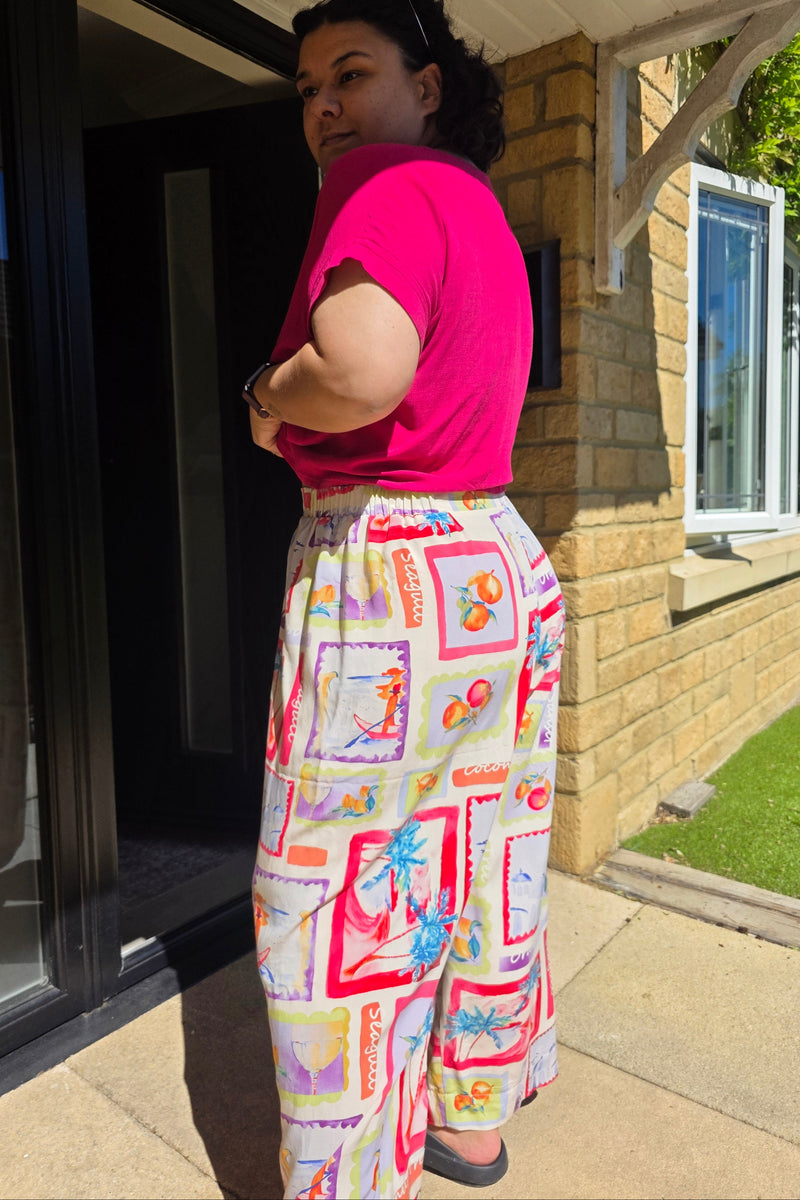 A person wearing a bright pink t-shirt and colorful patterned pants is lying on their back on a glass skylight in a backyard patio setting.