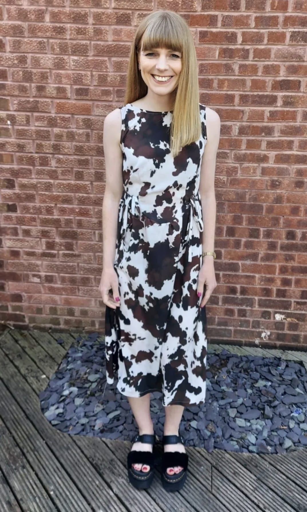 A woman with straight blonde hair and bangs stands outdoors in front of a red brick wall, wearing a sleeveless midi dress with a black, white, and brown cow print pattern. She is wearing black platform sandals and holding a small black handbag.